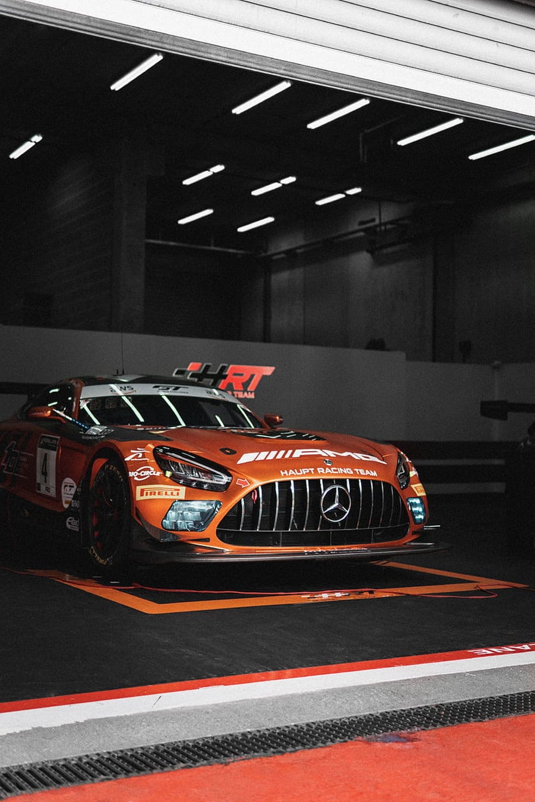 Orange Mercedes-AMG GT3 race car parked in a dark garage under bright fluorescent lights with Pirelli logos.