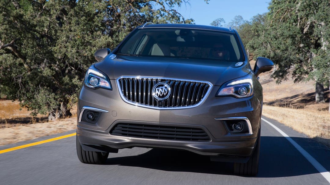 Buick Envision SUV driving on a sunlit road with green trees and golden hills in the background.