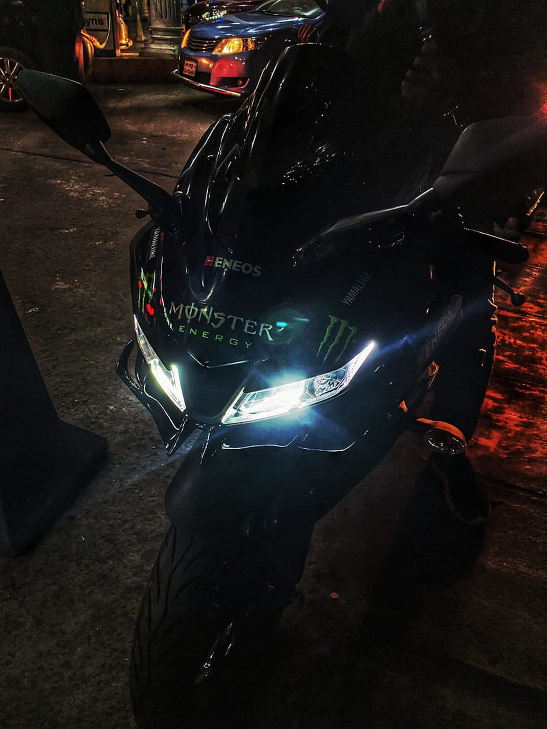 Yamaha R15 motorcycle with Monster Energy decals parked on wet pavement at night under bright LED headlights.