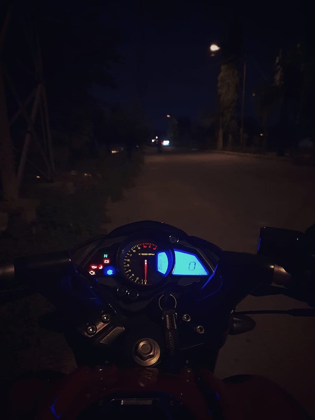 Motorcycle instrument cluster glowing blue at night with a needle tachometer and blurred streetlights behind.