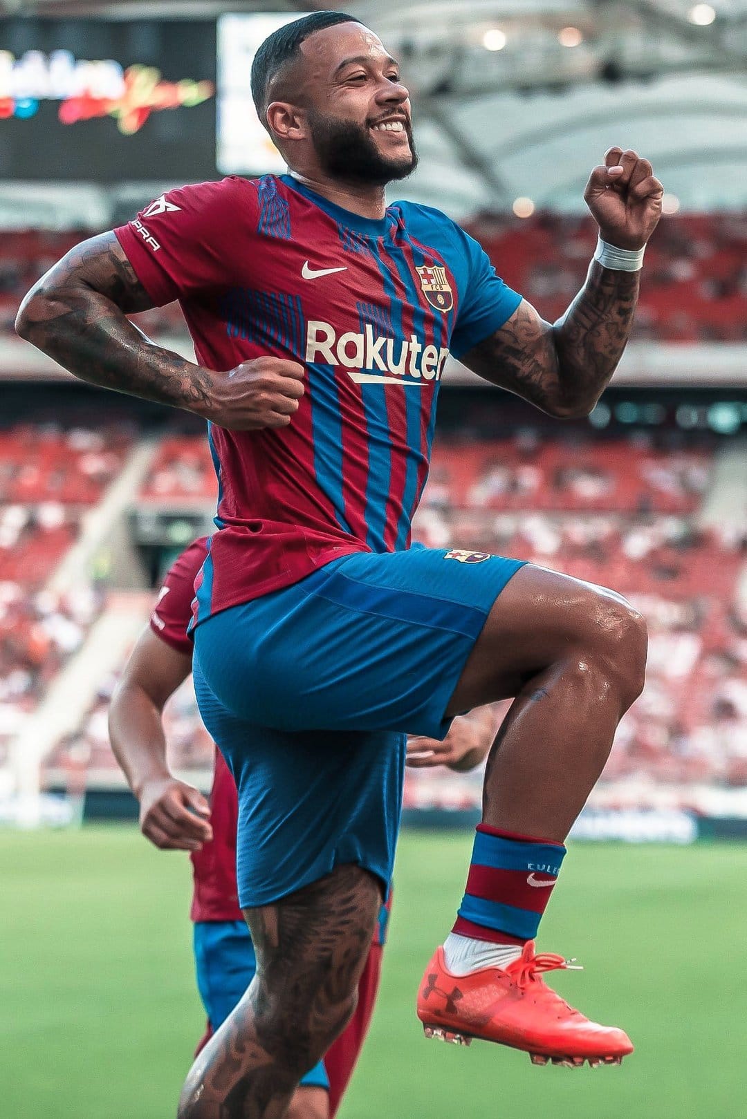 Memphis Depay celebrates in a Barcelona kit with a raised fist while wearing orange cleats on a grass pitch.