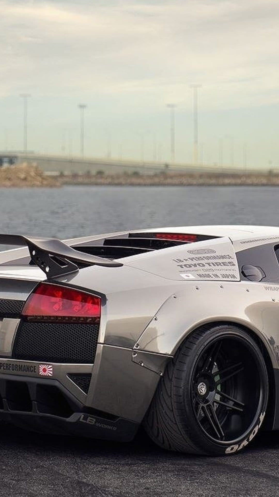 Lamborghini Murciélago with a widebody kit and rear wing parked at a hazy industrial port during sunset.