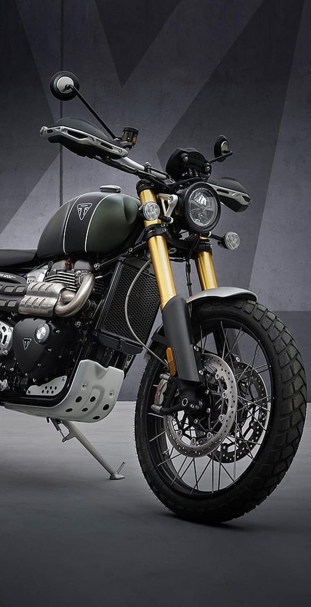 Triumph Scrambler motorcycle featuring gold front forks, knobby tires, and a green fuel tank on a grey surface.