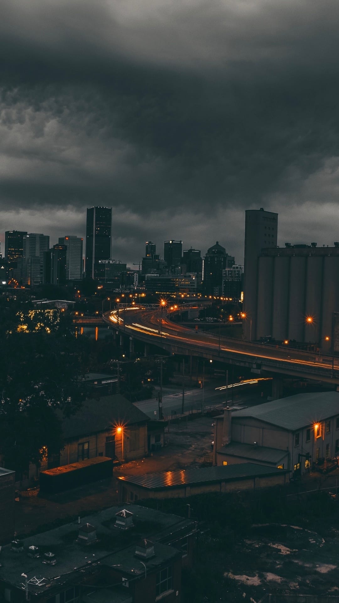 Glowing amber traffic trails on a winding urban overpass contrast against a dark, cloudy night city skyline.