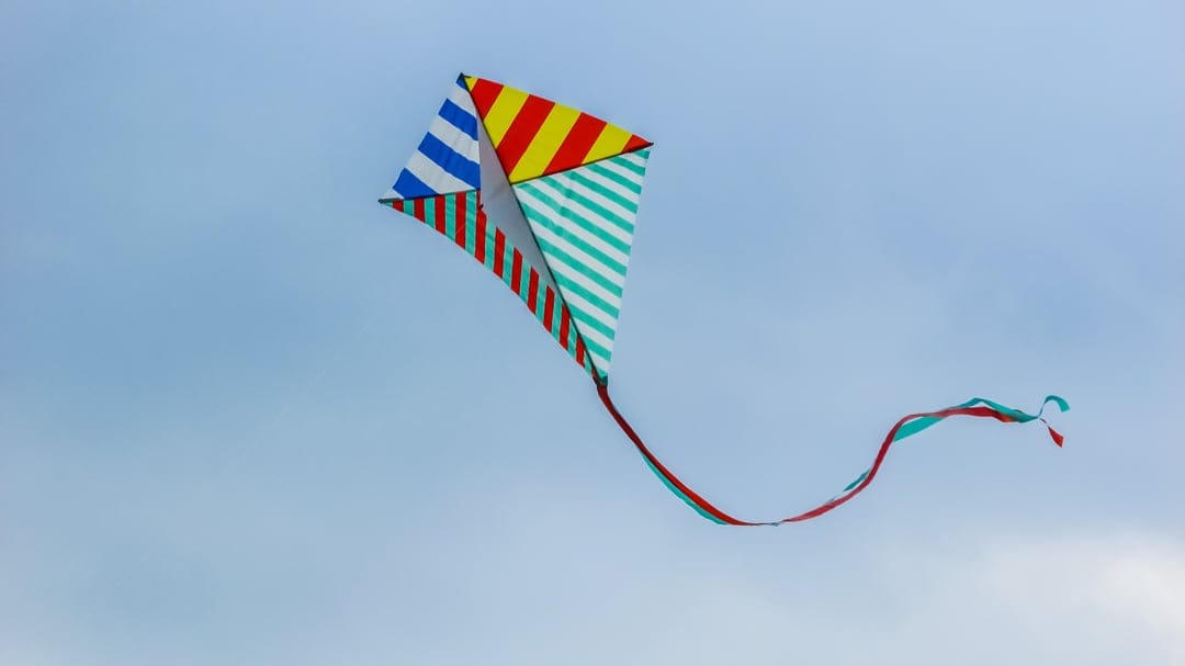 Multicolored striped diamond kite with red and turquoise ribbon tails soaring through a clear pale blue sky.