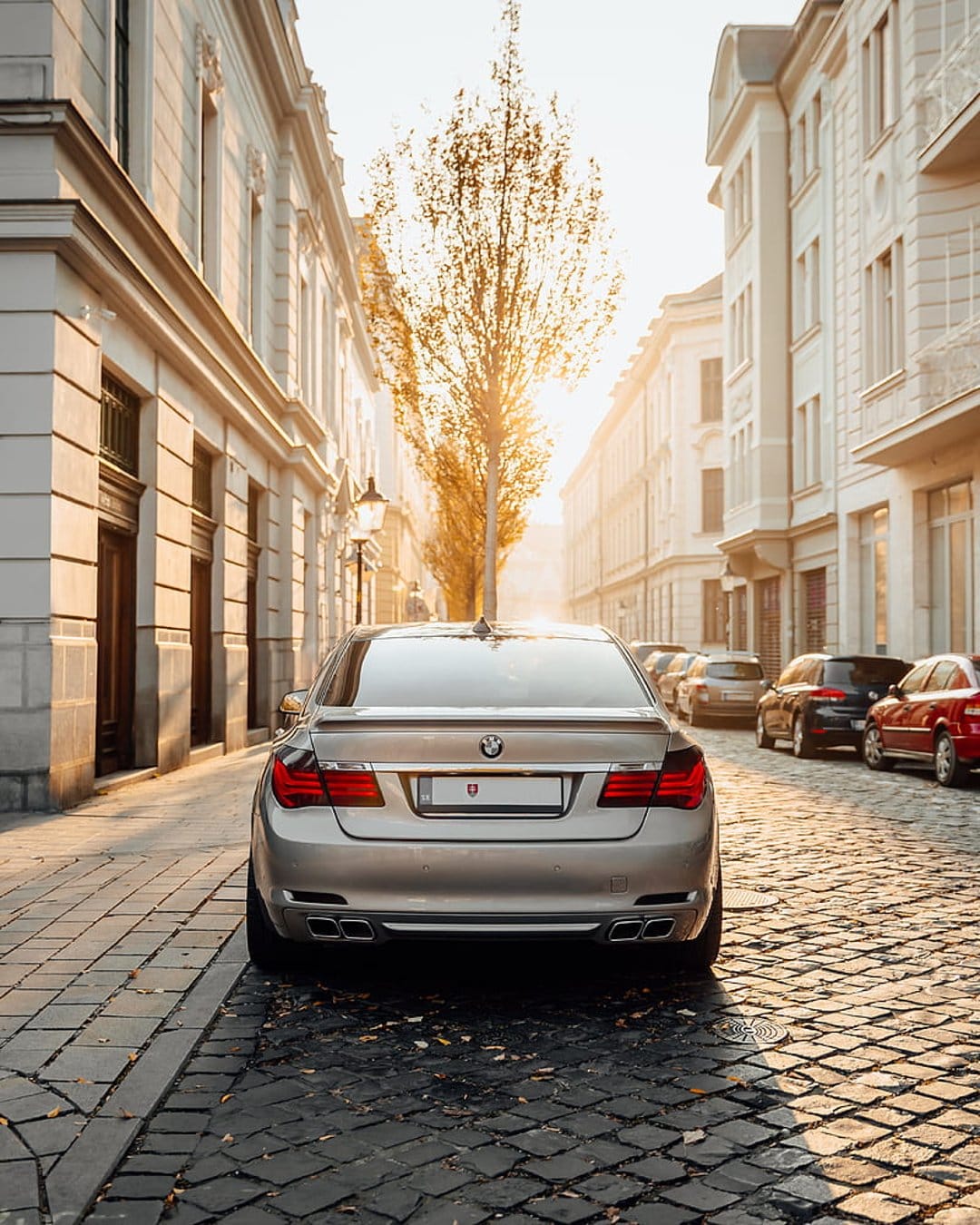 Silver BMW sedan parked on a cobblestone street with golden sunrise light filtering through autumn tree branches.
