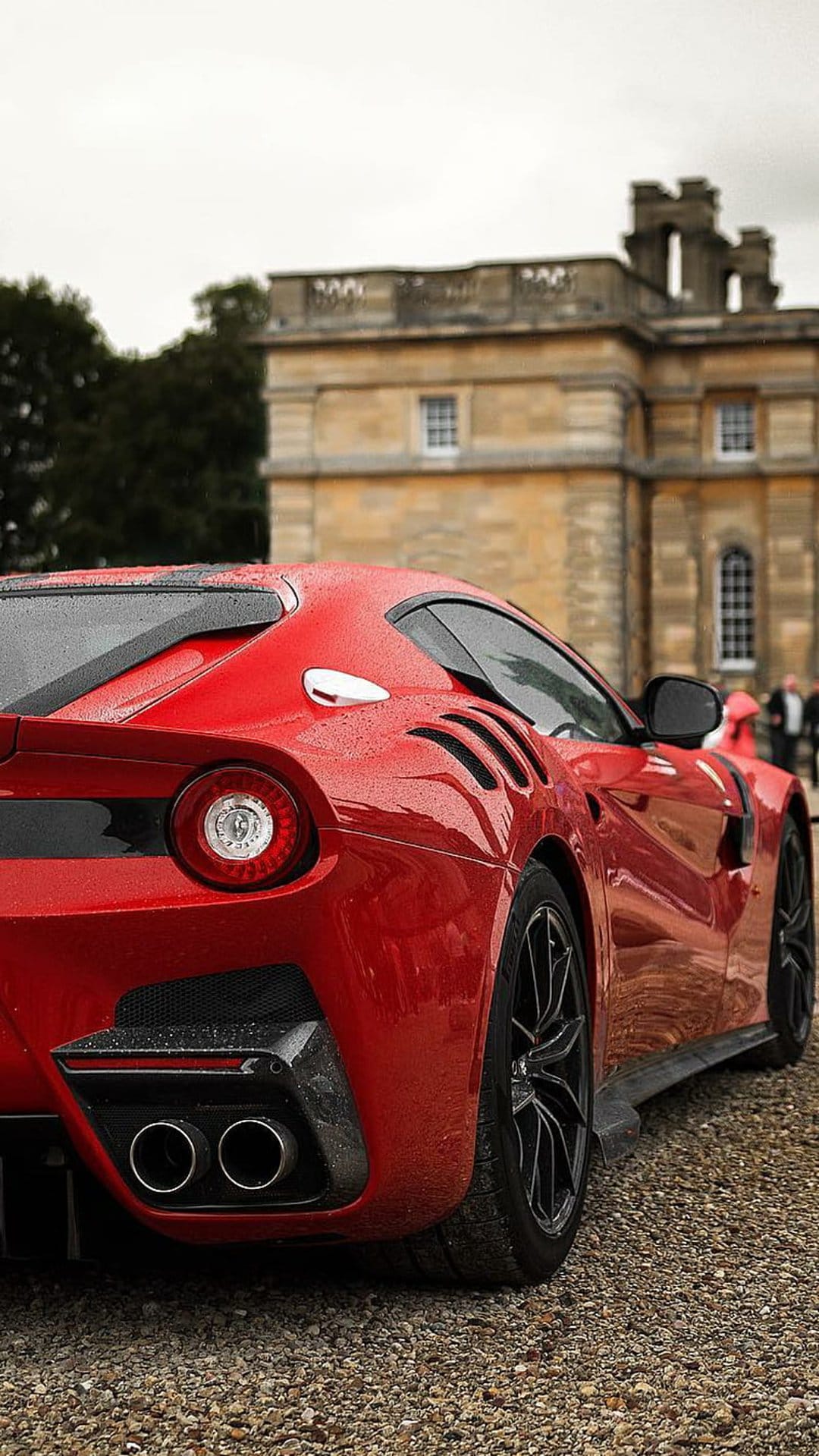 Ferrari F12 sports car with wet red paint parked on a gravel path outside a historic English country estate.