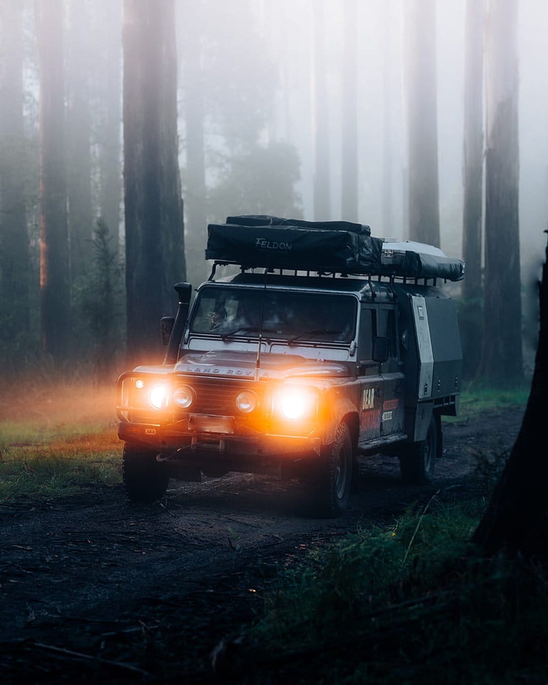 Land Rover Defender with a rooftop tent drives through a dense foggy forest with glowing headlights on a dirt road.