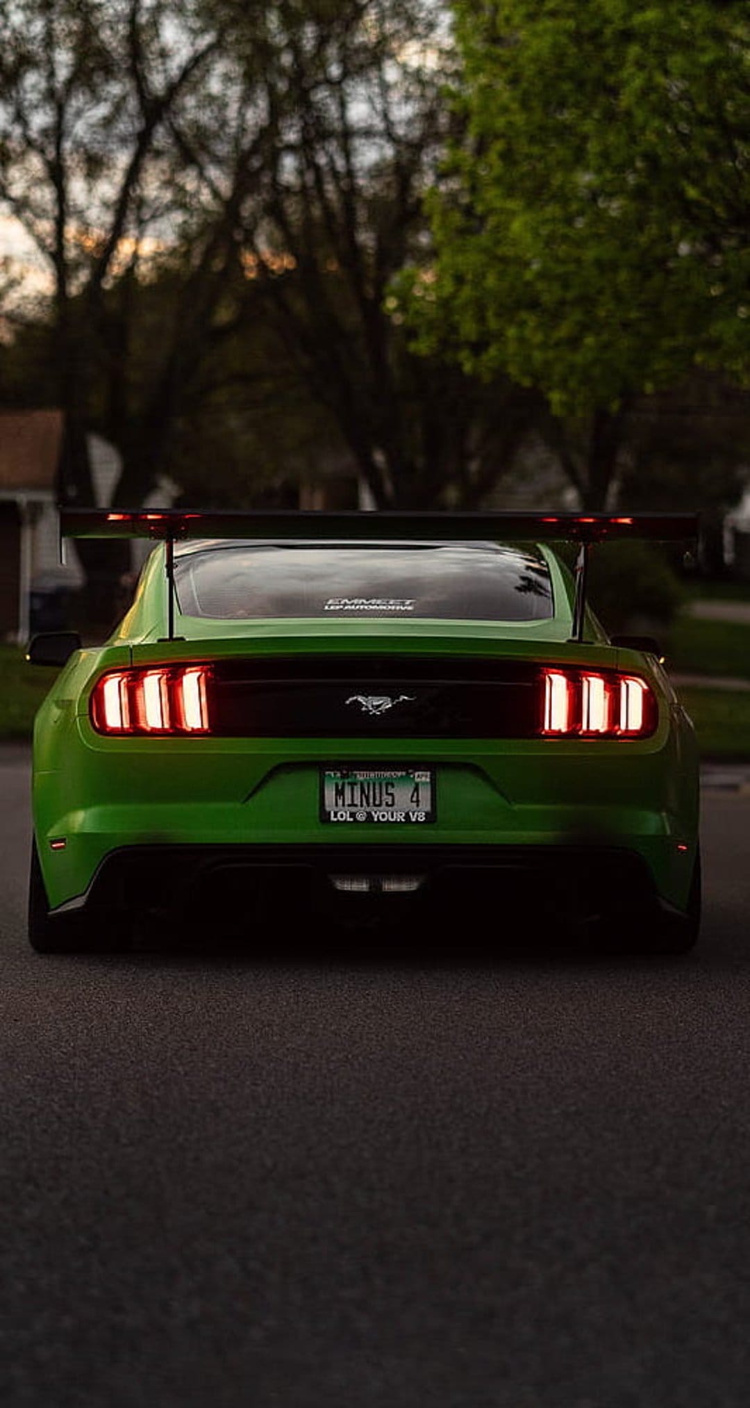 Green Ford Mustang rear view with a massive black spoiler and glowing red taillights on an asphalt road.