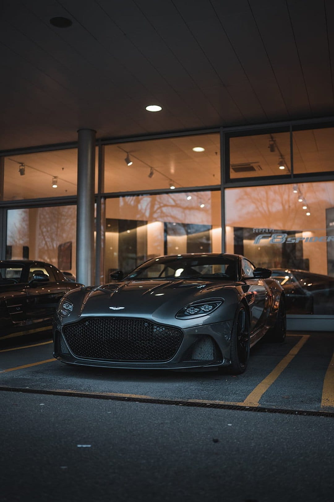 Dark grey Aston Martin DBS Superleggera with a large grille reflects warm lights inside a modern car showroom.