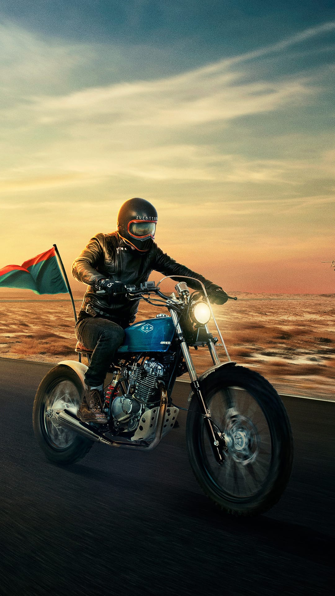 Biker in a leather jacket riding a blue vintage motorcycle with a trailing flag against a desert sunset sky.