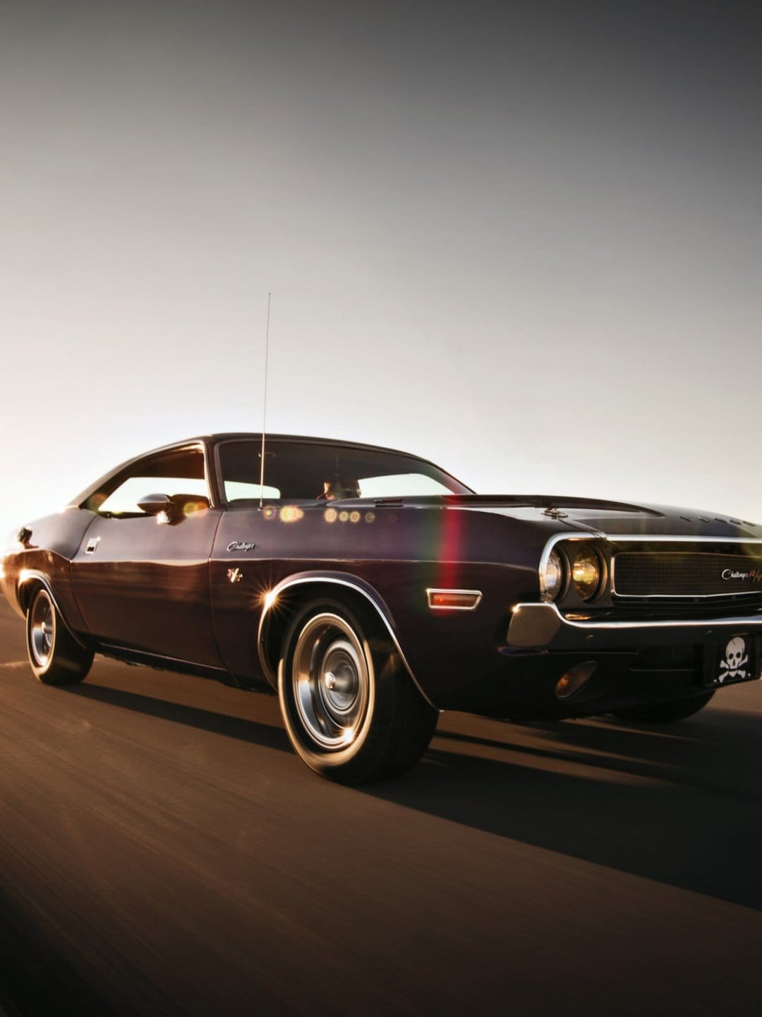 Purple classic muscle car driving on a coastal road at sunset with golden lens flares and motion blur details.