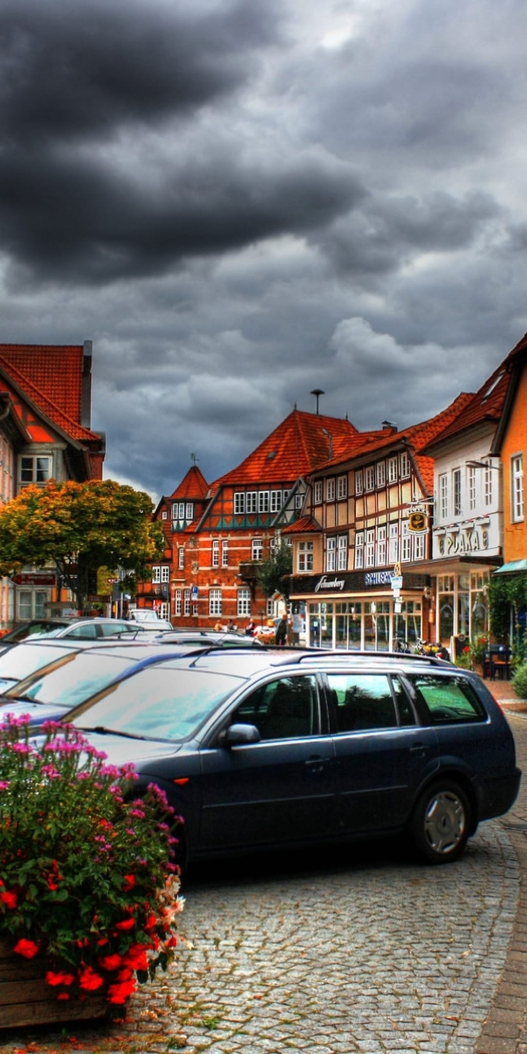 Weathered cobblestone street lined with half-timbered buildings and red flowers under a dark, stormy sky.