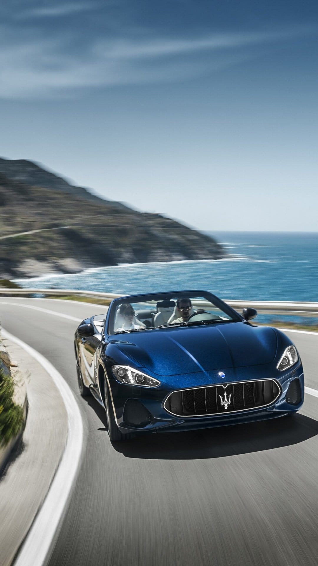 Deep blue Maserati convertible driving on a winding coastal road overlooking the ocean with motion blur.