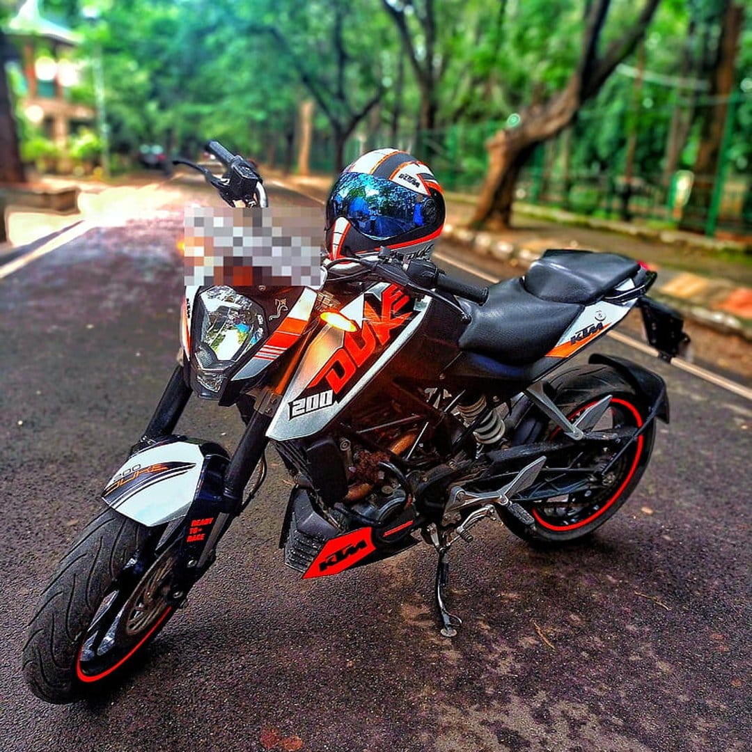 KTM Duke 200 motorcycle with a reflective helmet on the seat, parked on an asphalt road near green trees.