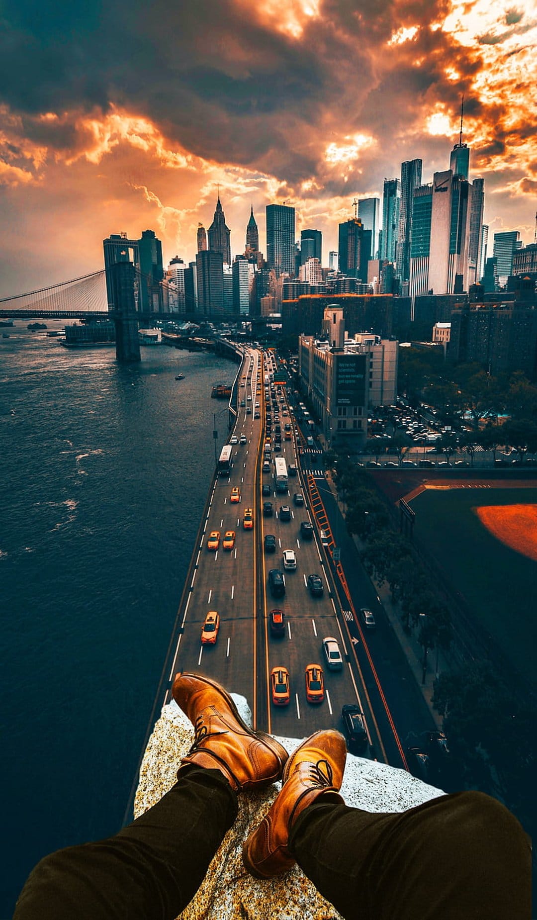 Man's legs in brown boots dangling over a highway and New York City skyline under a fiery sunset sky.