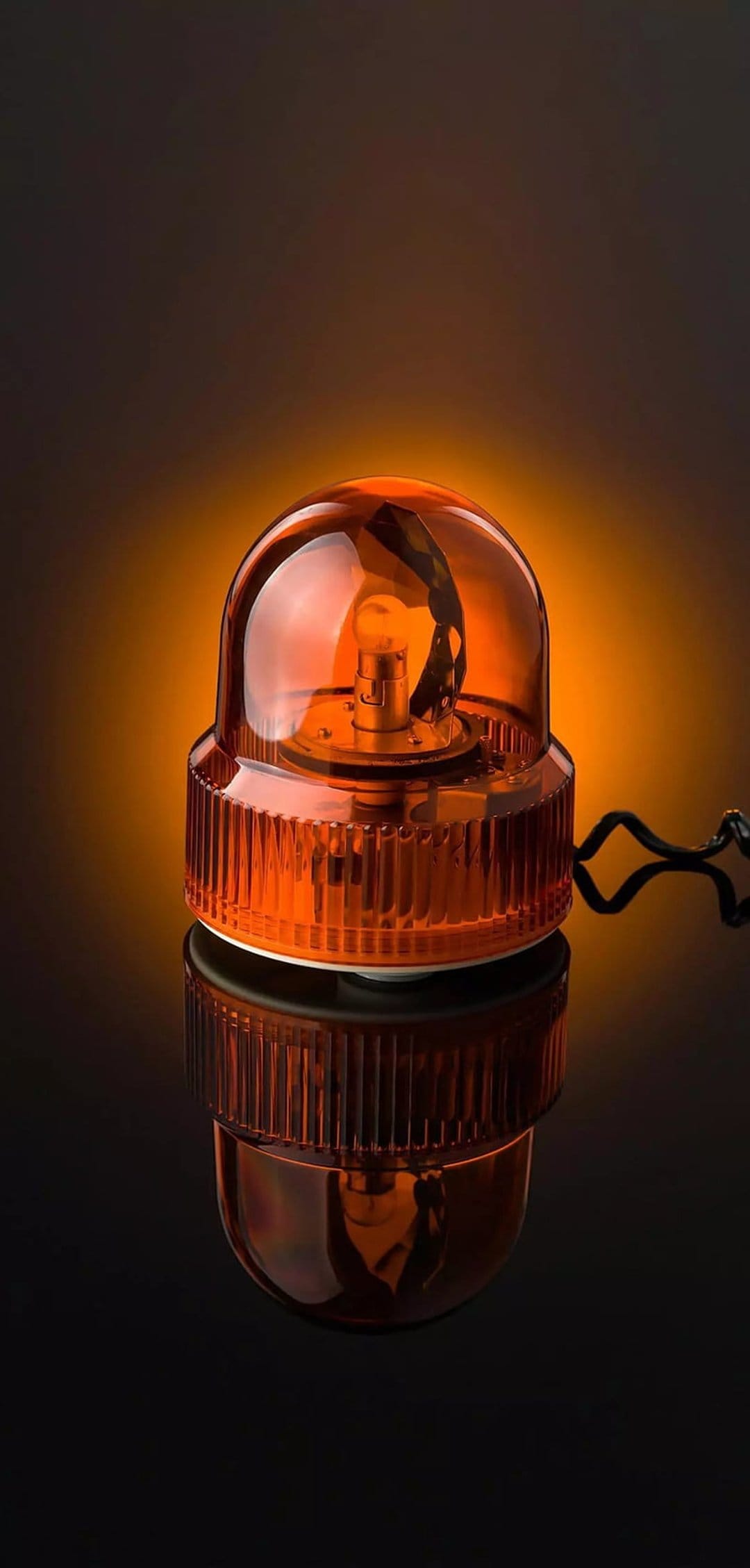 Orange emergency beacon with a ribbed amber dome reflects off a glossy black floor in a dark studio setting.