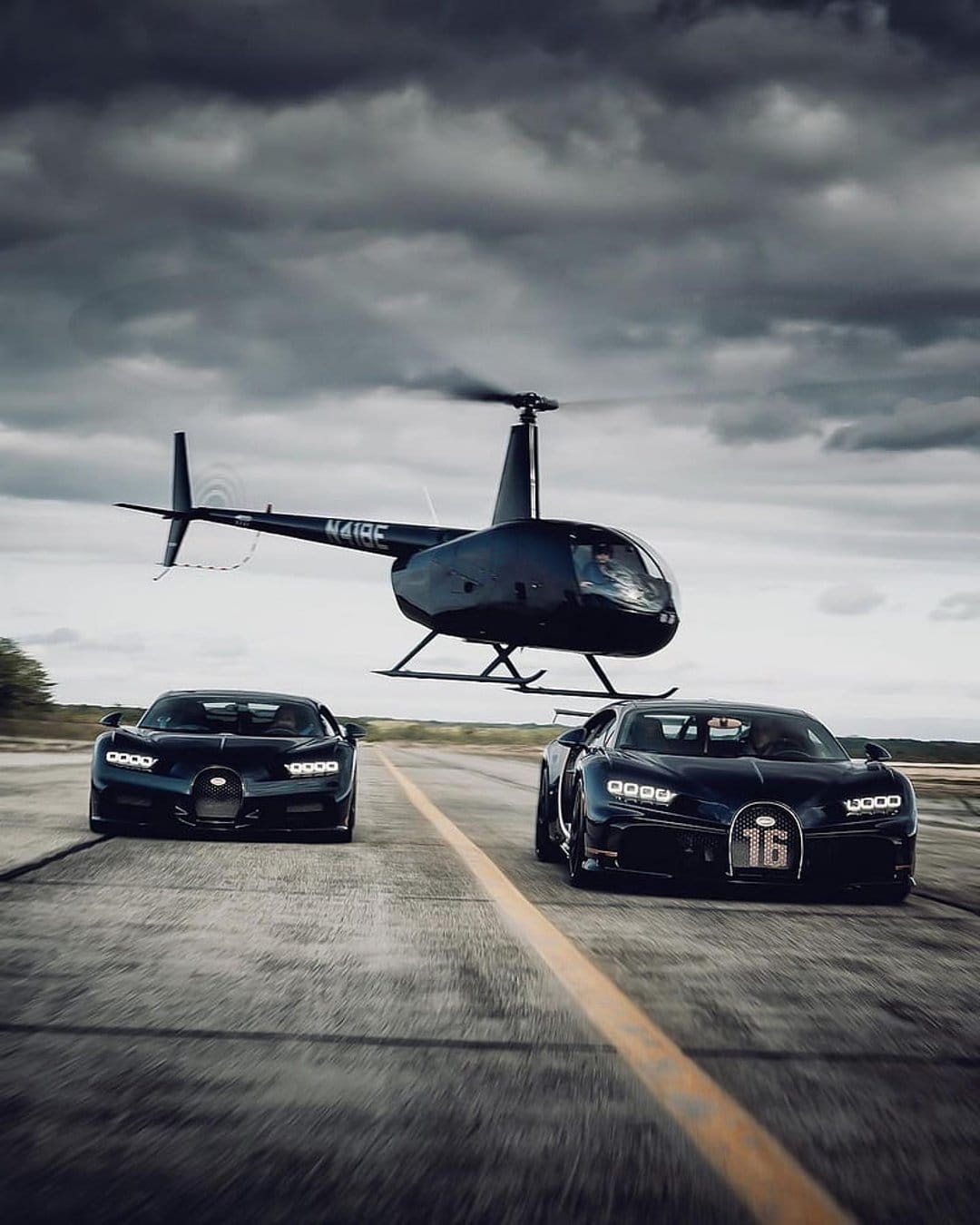Black Bugatti hypercars speed along a runway beneath a low-flying helicopter under a dark, stormy sky.
