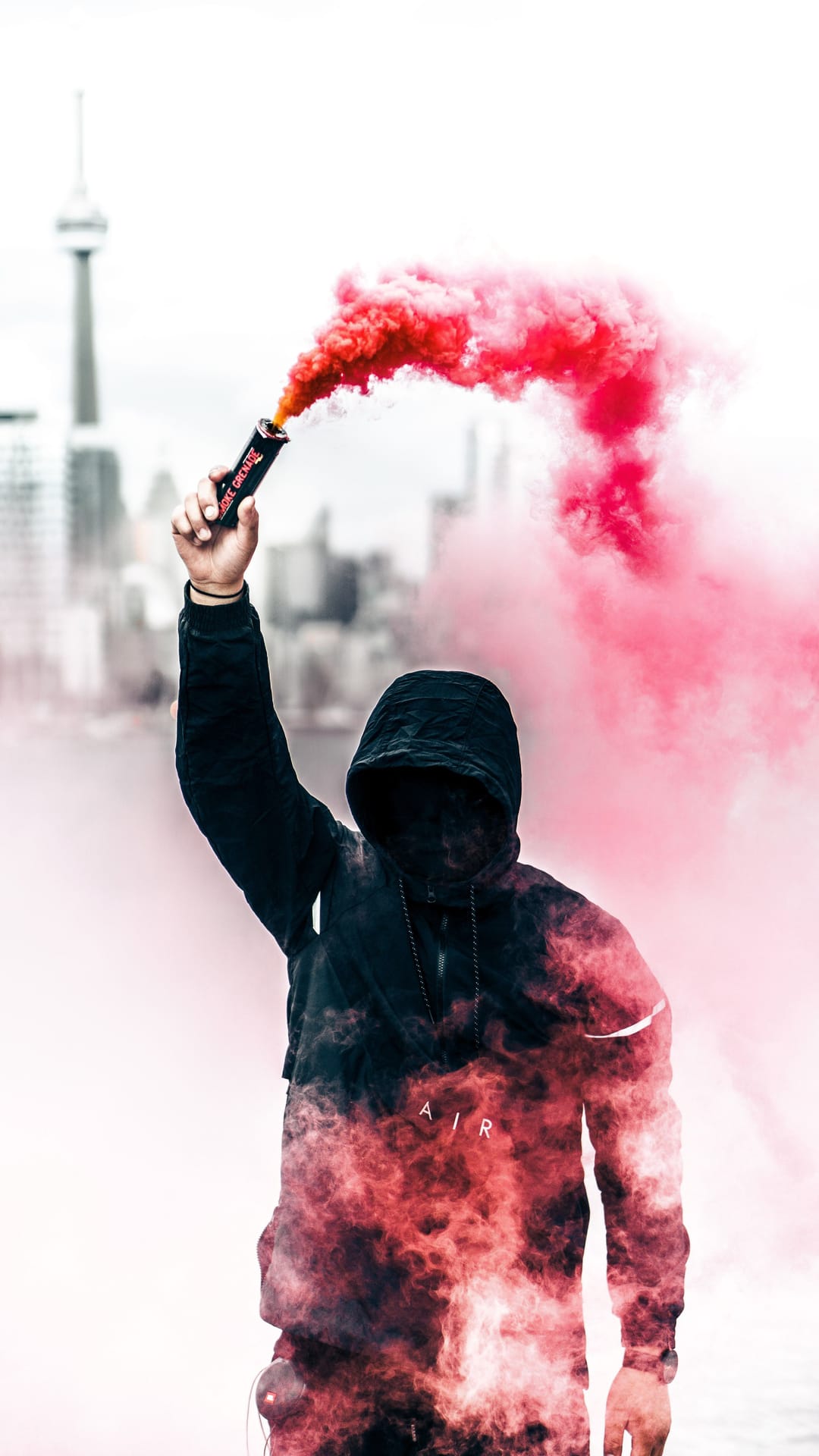 Hooded person holds a burning red smoke flare with a blurred city tower and skyscrapers in the background.
