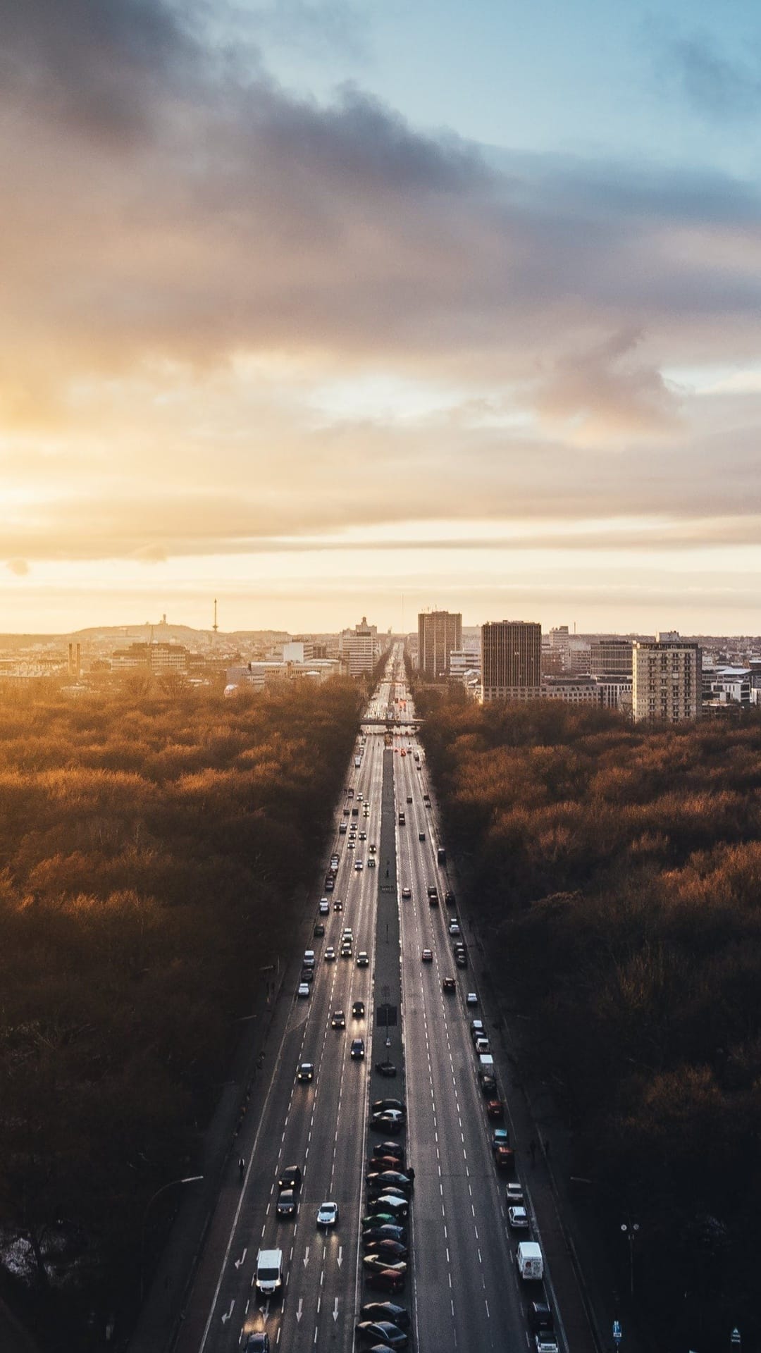 Multi-lane highway with car lights cutting through orange autumn forest toward a hazy sunset city skyline.