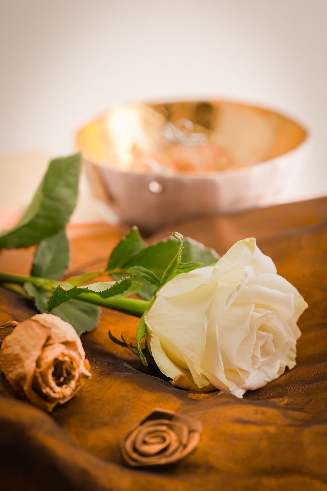 White rose and dried flower lie on brown satin next to a small bowl of rings in a romantic still life scene.