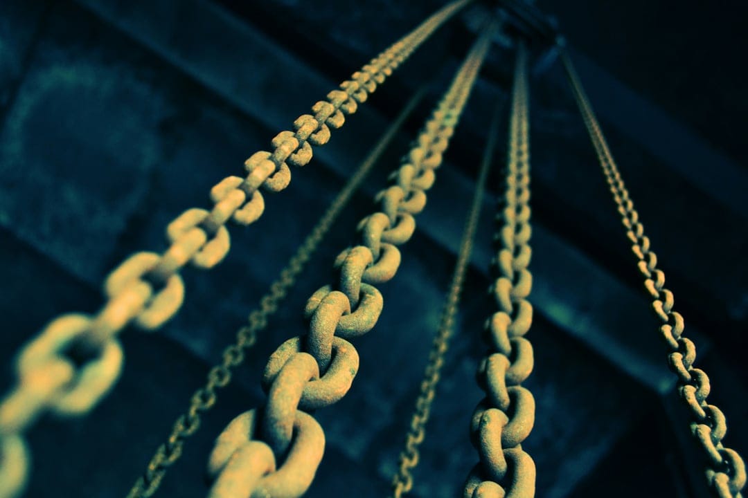 Rusty iron chains with heavy weathered links hang vertically from above against a dark, moody backdrop.