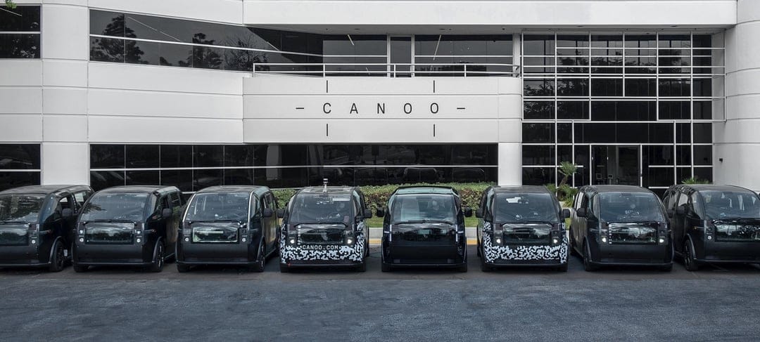 Line of pod-shaped Canoo electric vehicles in black and camouflage parked at a white modern building.