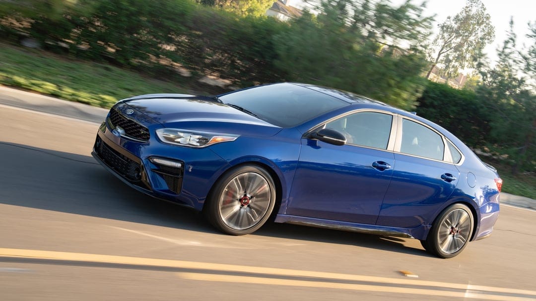 Blue Kia Forte GT driving along a suburban road with motion blurred greenery and rotating alloy wheels.