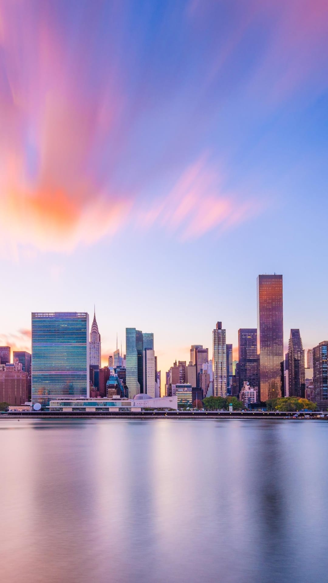 Modern city skyline silhouettes under a pink and orange sunrise with clouds reflecting in the still water.
