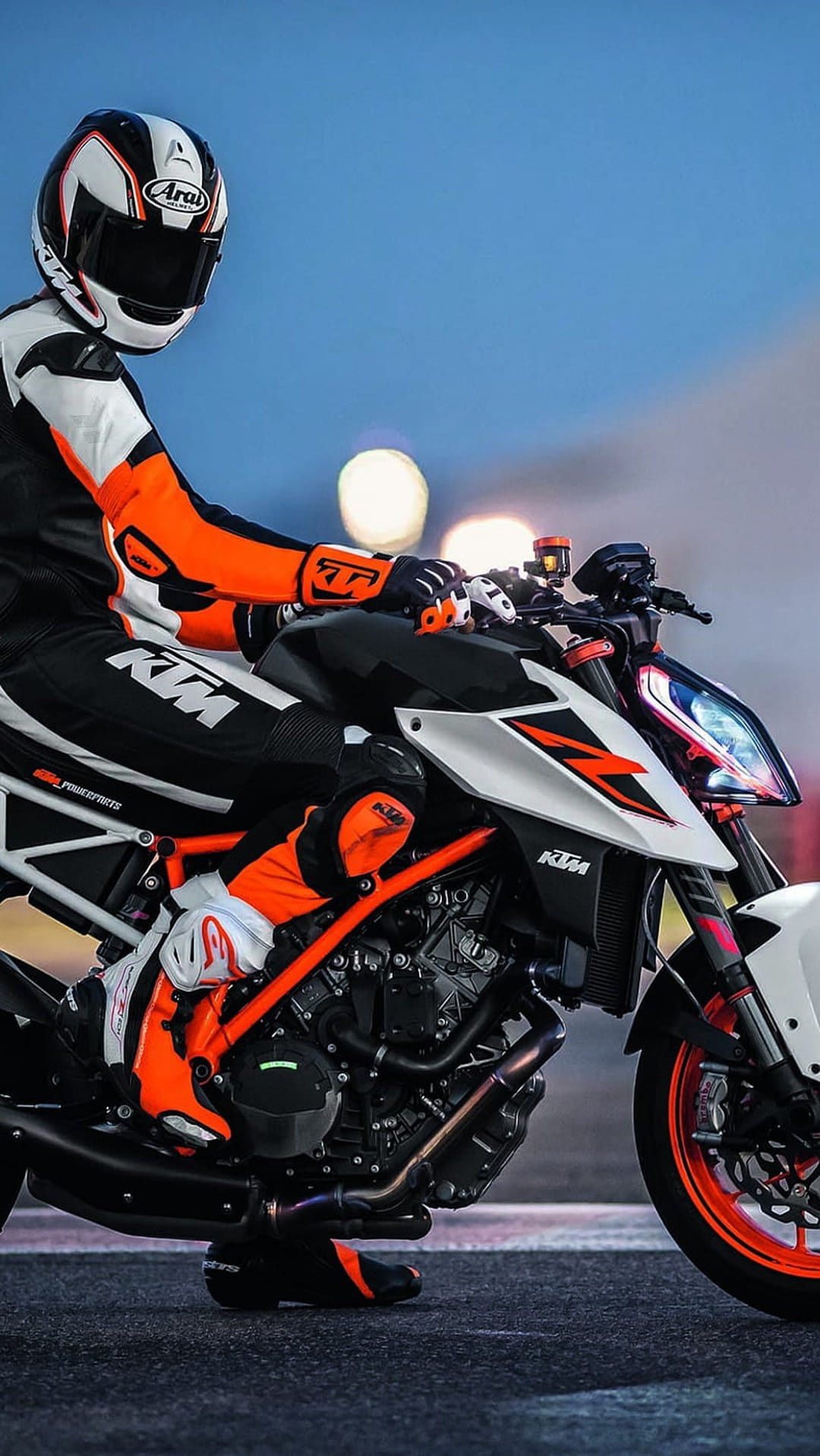 Motorcyclist in orange and white racing leathers sits on a sportbike with bokeh lights in the background.