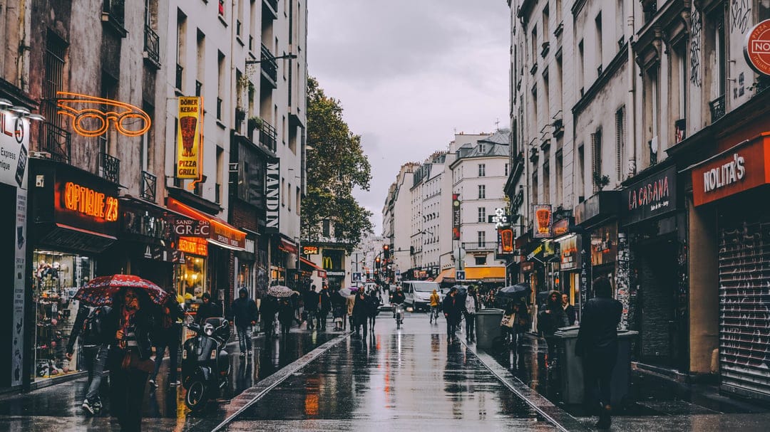 Silhouetted people with umbrellas walk past glowing shops on a wet cobblestone street in Paris at dusk.
