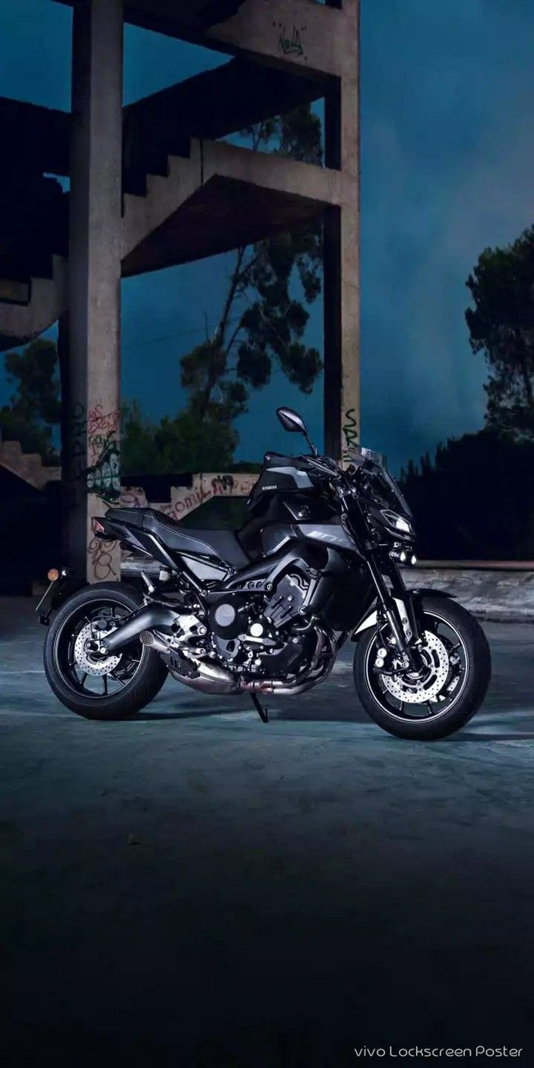Black Yamaha motorcycle parked in a concrete brutalist ruin with graffiti under a dark blue twilight sky.