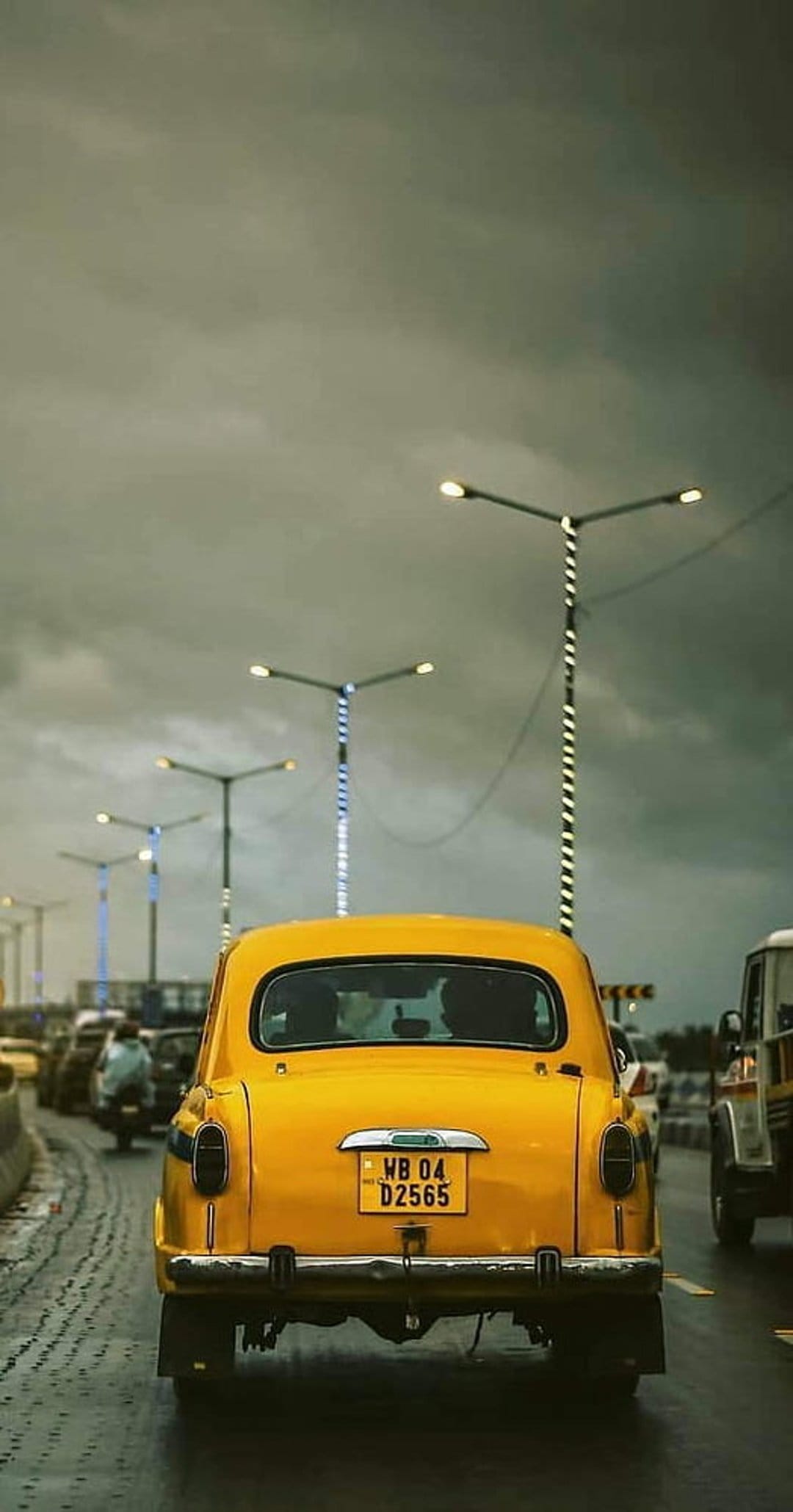 Yellow vintage taxi driving on a wet asphalt road at dusk with glowing streetlights and city traffic background.