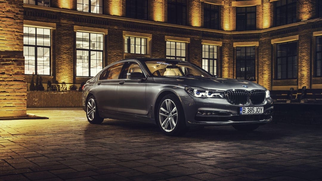 Charcoal grey BMW sedan with glowing headlights parked on cobblestones in front of a lit brick building at night.