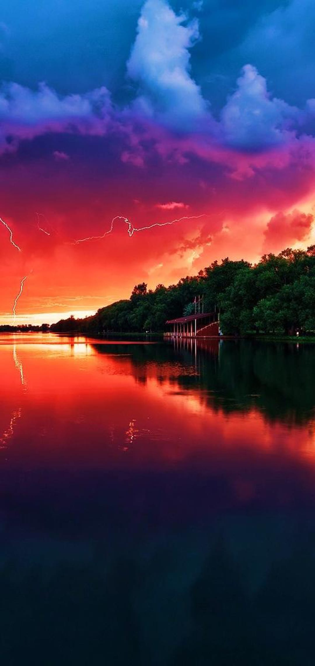 Lightning bolts strike over a crimson and violet sunset sky reflected in a calm forest lake with a small cabin.