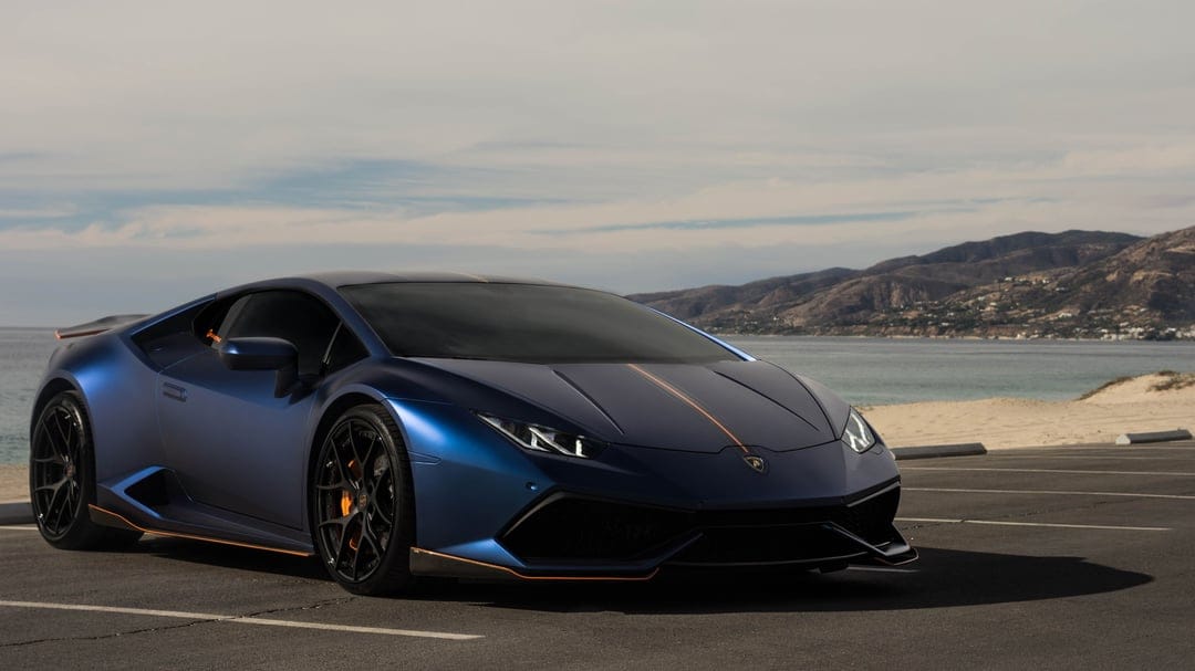 Blue Lamborghini Huracan with orange accents parked on a coastal road against a backdrop of ocean and mountains.