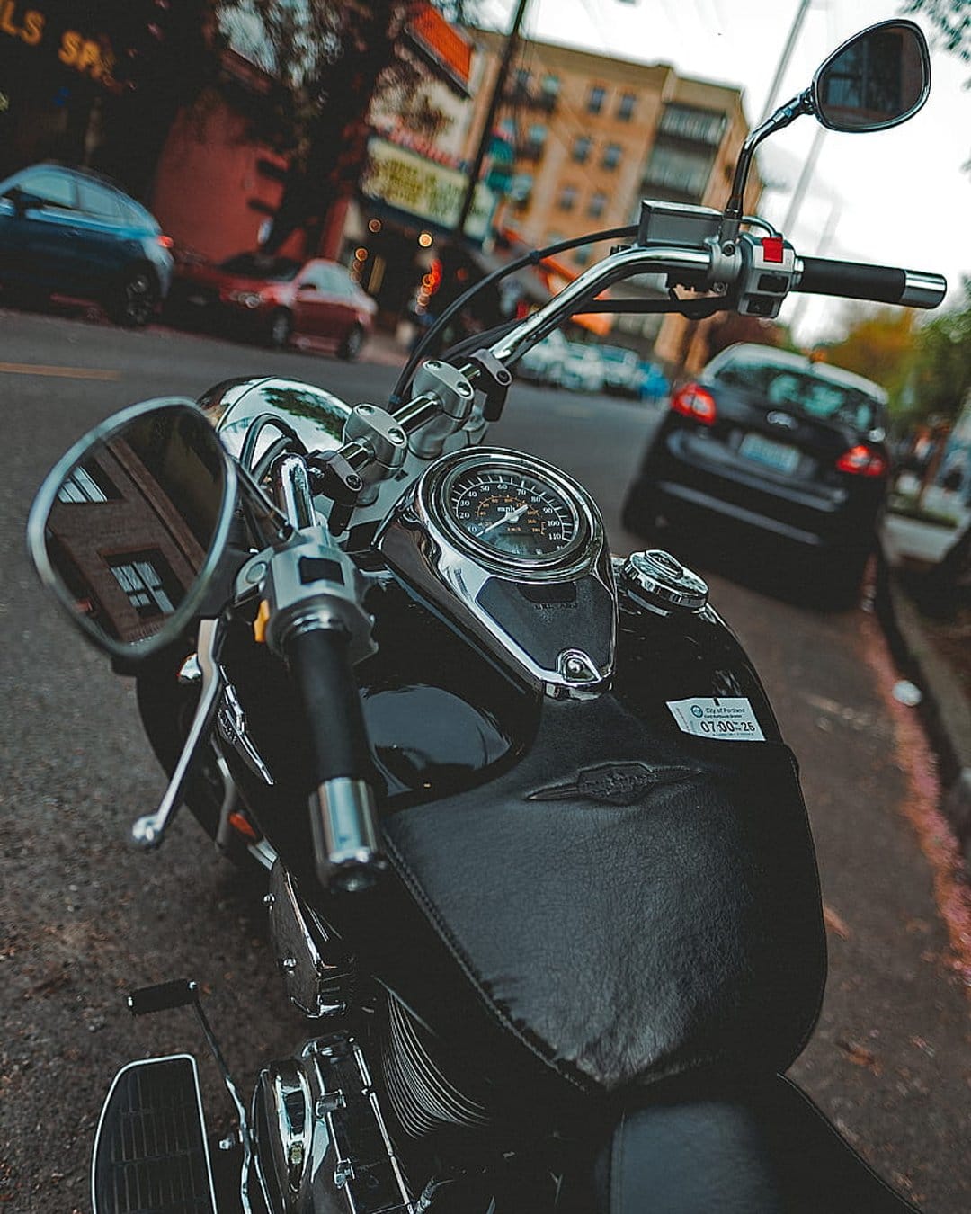 Black motorcycle handlebars and speedometer on a city street with a blurred urban background and asphalt road.