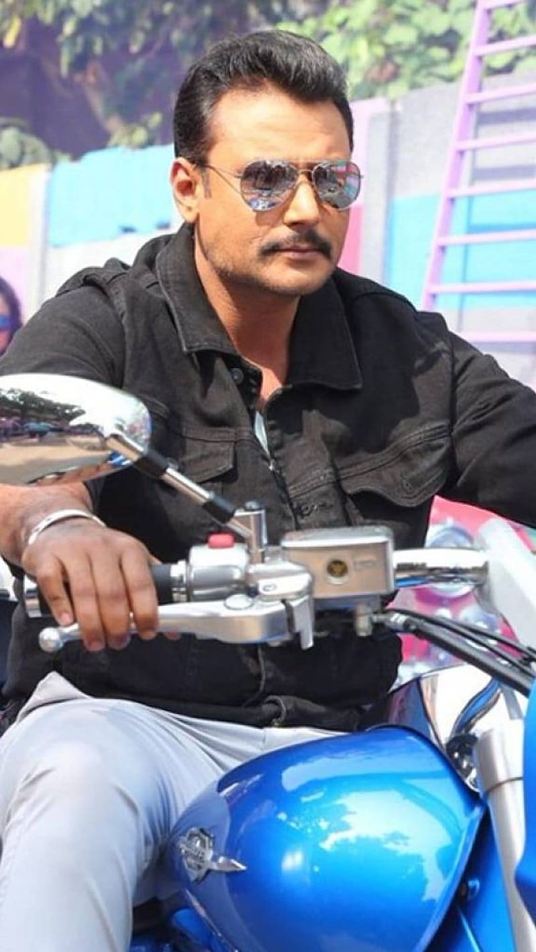 Actor Darshan riding a blue motorcycle wearing a black denim jacket and sunglasses in a vertical portrait.