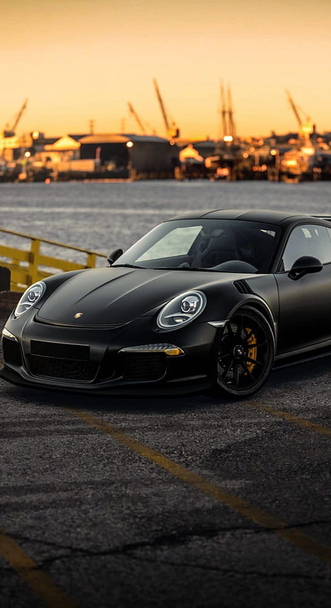 Matte black Porsche 911 GT3 with yellow calipers at a dockside sunset with industrial cranes in the background.