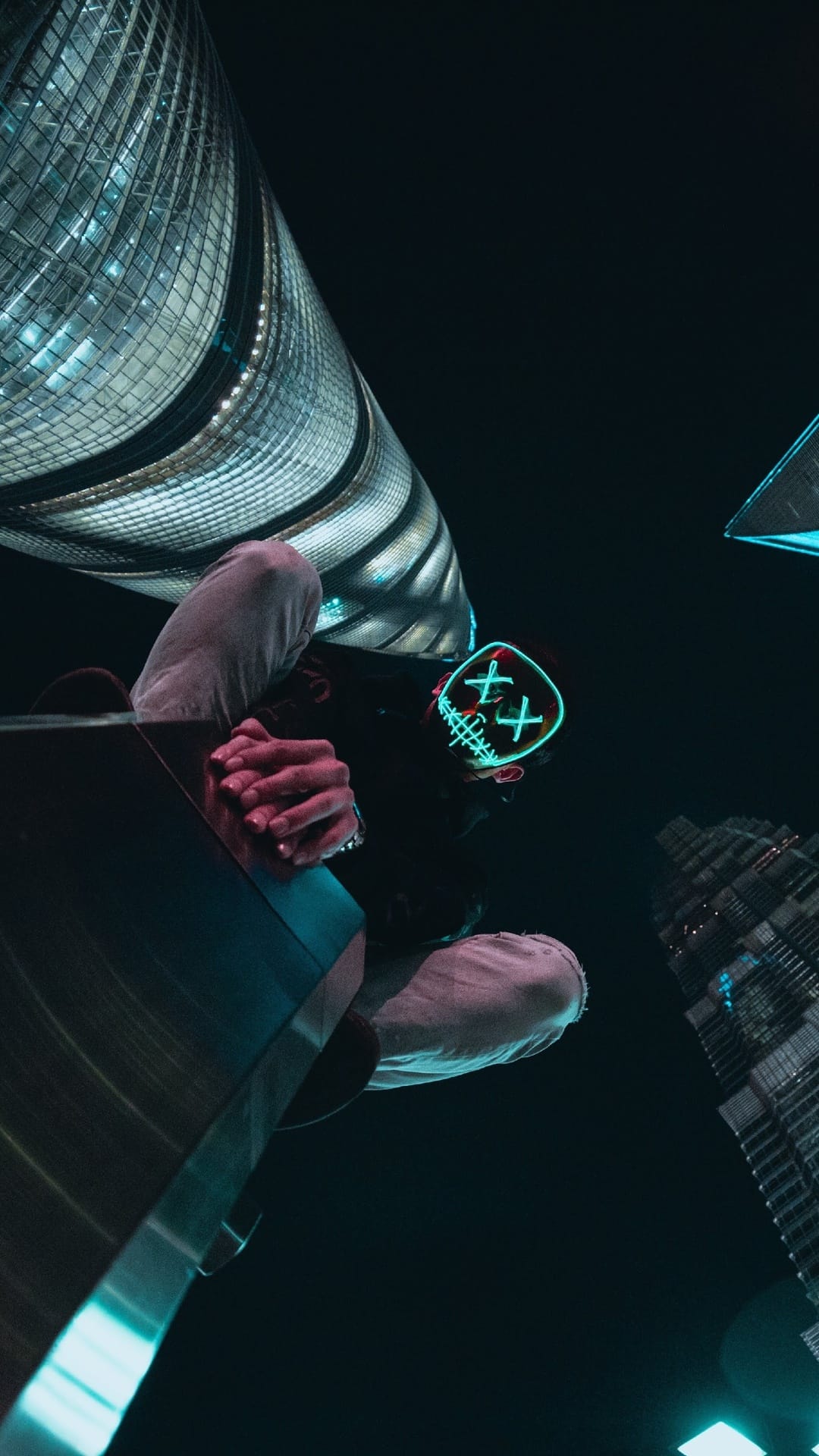 Masked figure with glowing green X eyes gazes down from high-rise skyscrapers against a dark urban night sky.