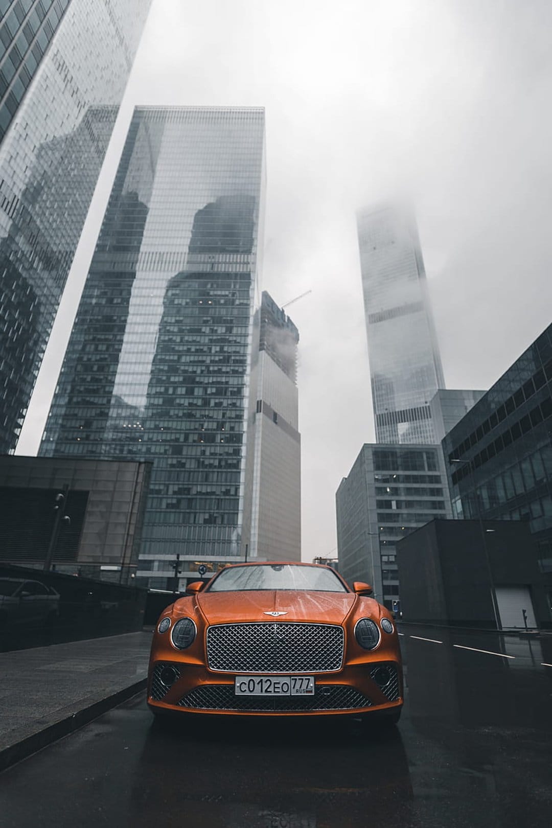 Orange Bentley Continental GT on a wet street surrounded by foggy skyscrapers in a vertical urban scene.