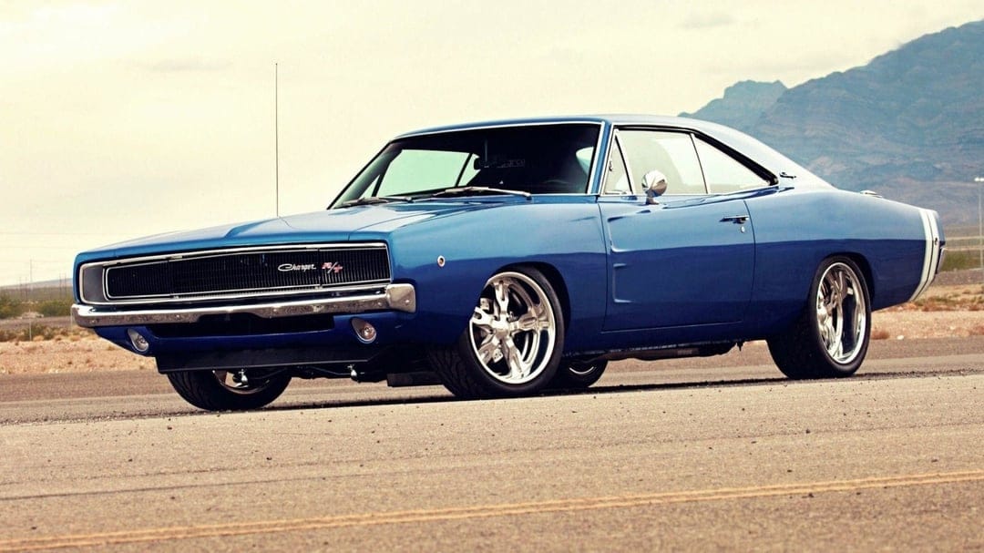 Blue 1968 Dodge Charger R/T with chrome wheels on an asphalt road against distant mountains and a pale sky.