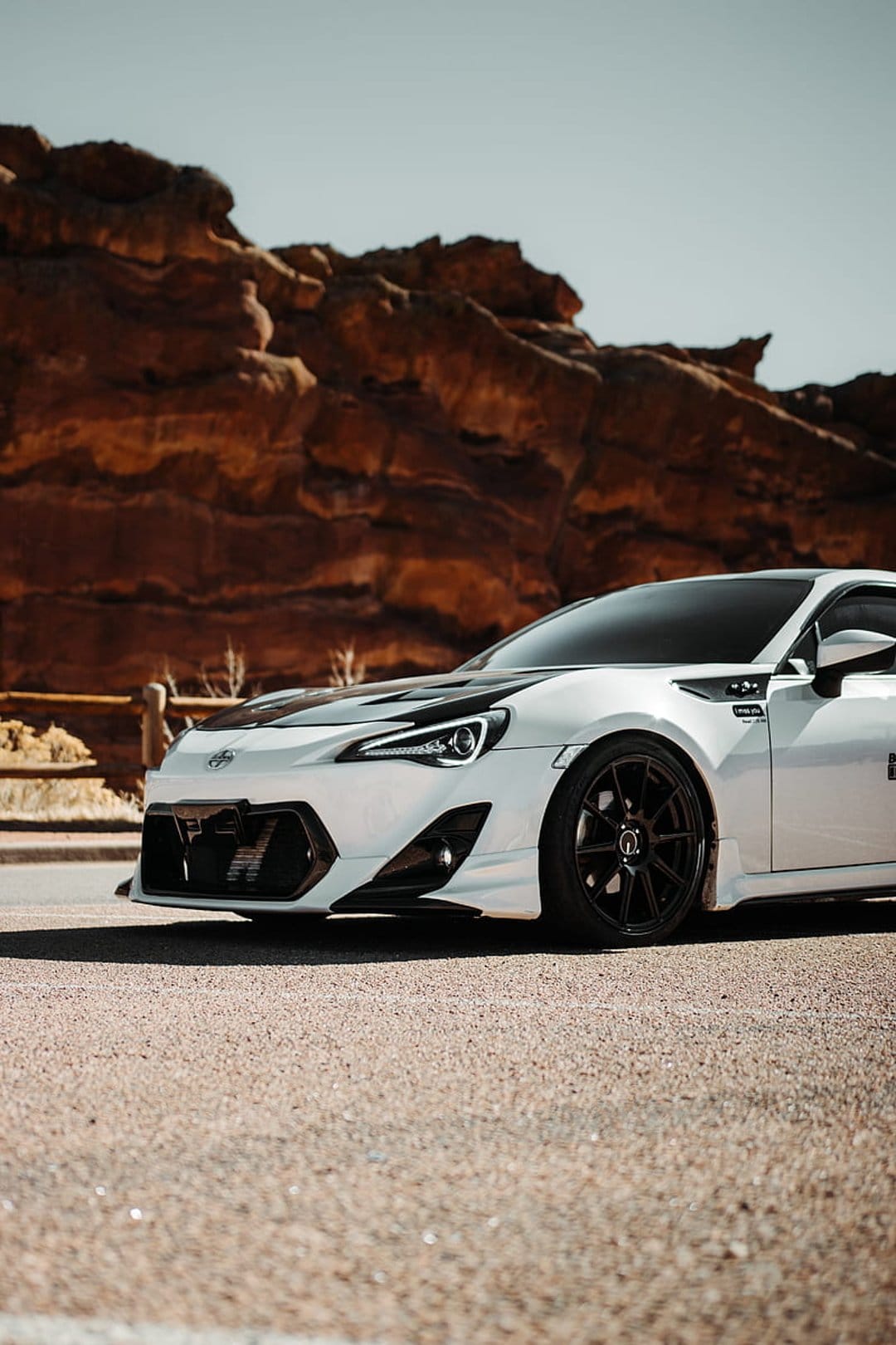 White Toyota GT86 sports car with a black hood on a desert road near massive red rock sandstone formations.