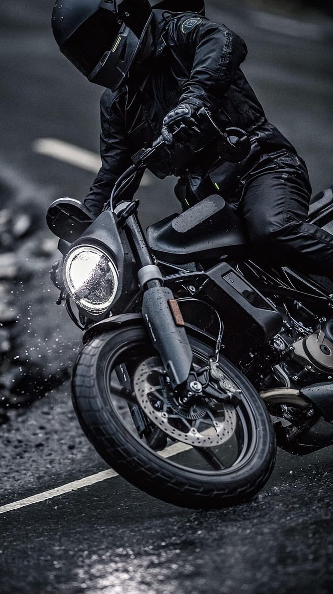Motorcyclist in black gear rides a dark sportbike on a wet road with glowing headlights and water spray.