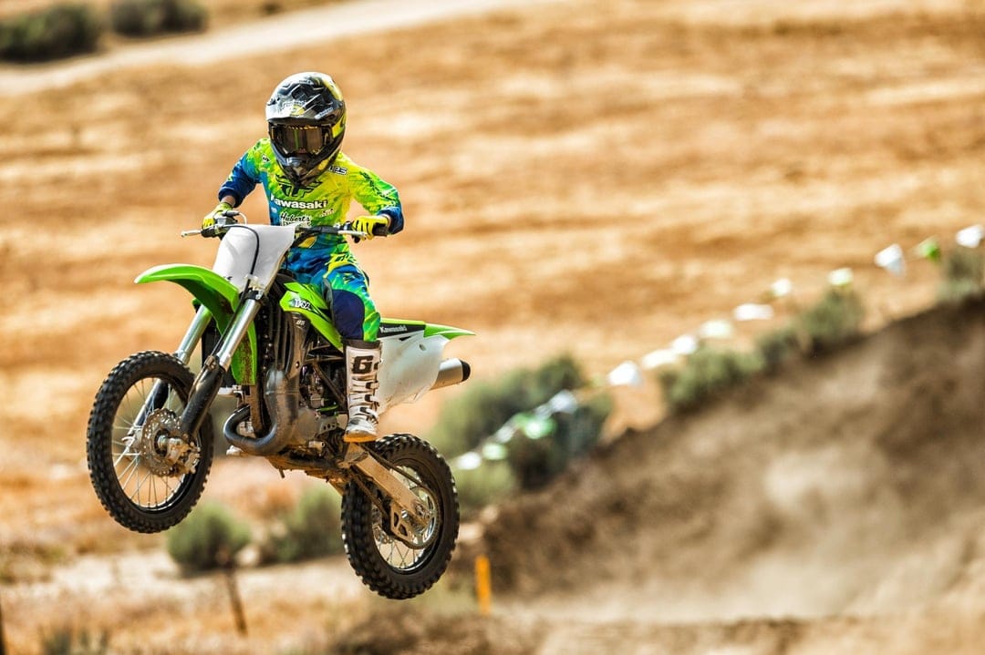 Motocross rider jumps a green Kawasaki dirt bike over a sunlit sandy track with blurred desert hills in the distance.