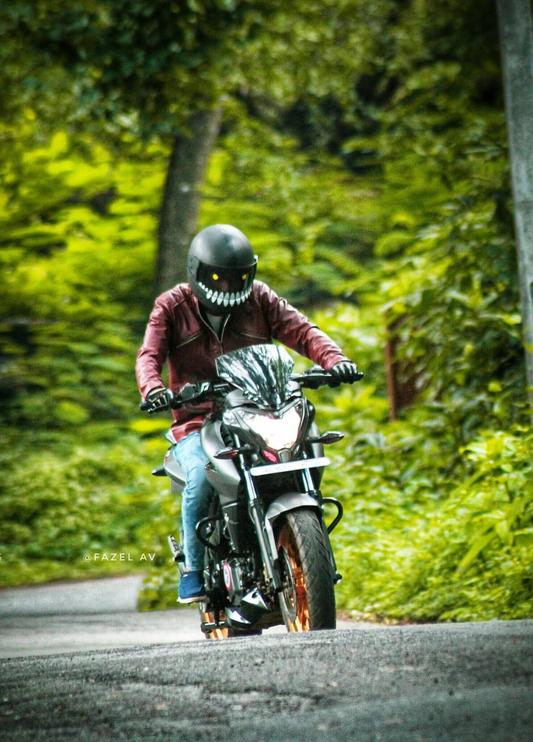 Motorcyclist in a monster helmet with glowing yellow eyes and a red jacket rides on a green forest road.