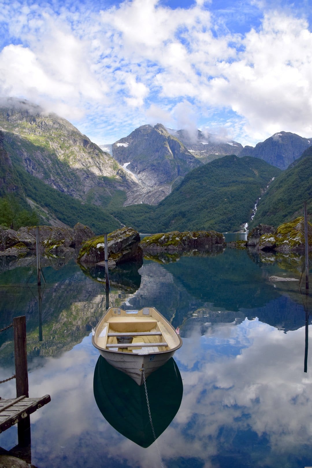 Small boat on a glassy lake reflecting steep green mountains and white clouds under a clear blue sky in Norway.