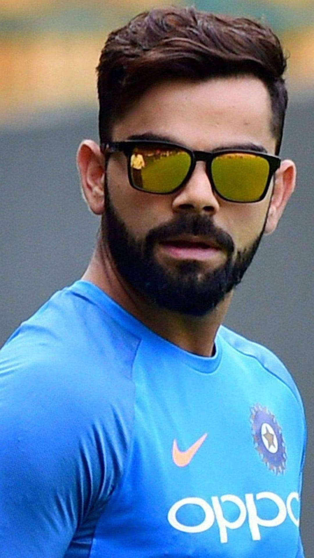 Virat Kohli wears gold reflective sunglasses and a blue Indian team jersey with his hair styled upwards.