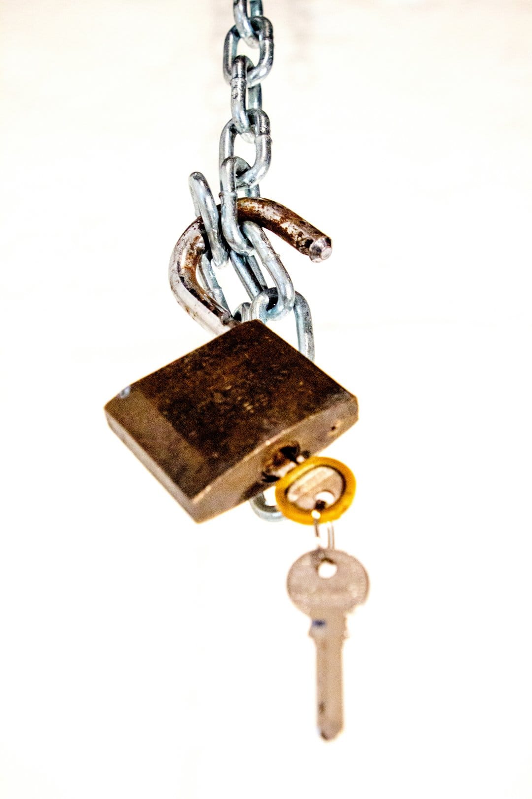 Brass padlock with a silver key inserted hangs from a heavy metal chain against a clean white background.
