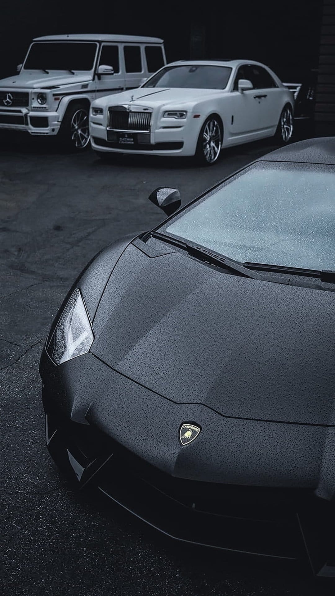 Lamborghini, Rolls-Royce Ghost, and G-Wagon parked on wet pavement with raindrops reflecting cinematic light.